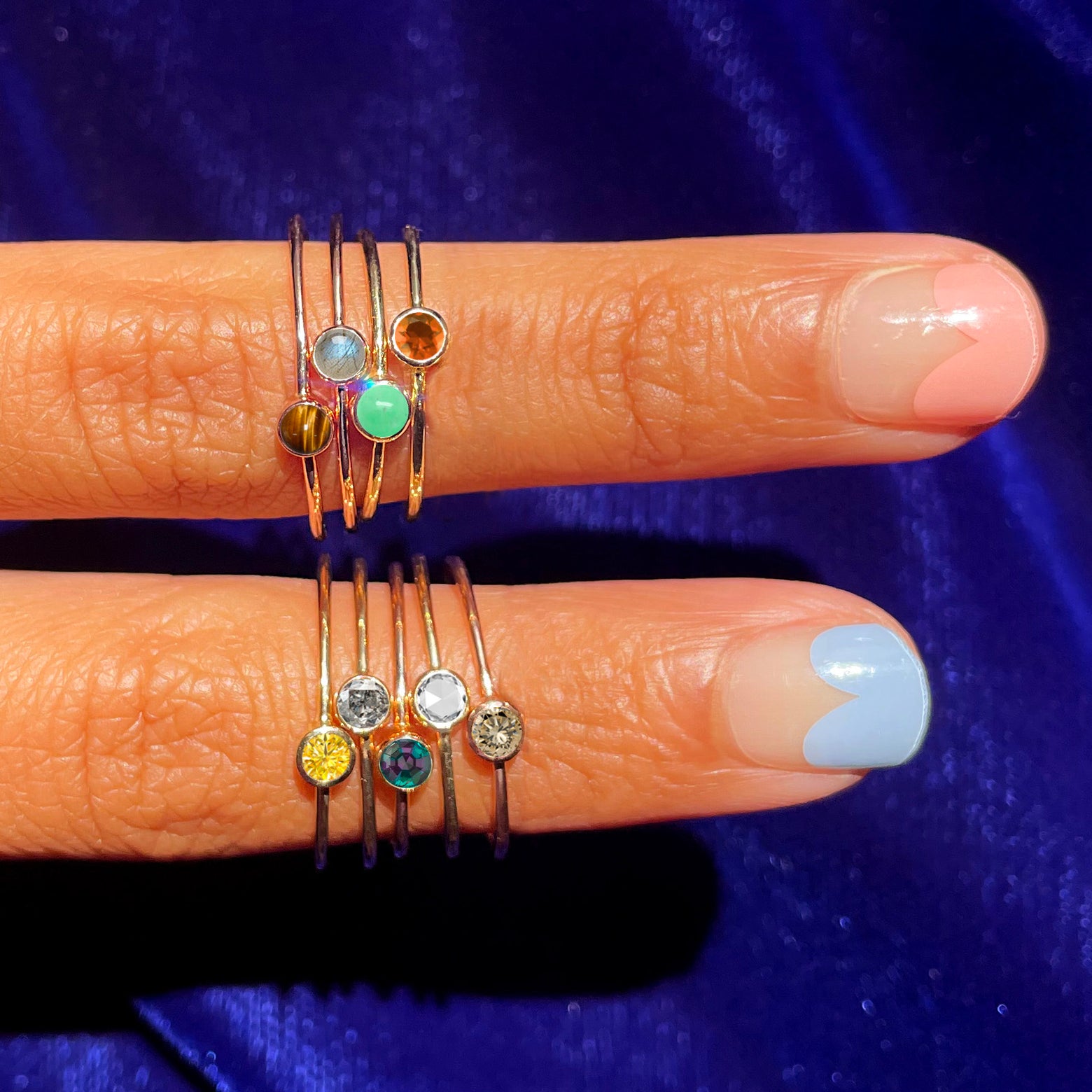 Two fingers with colorful nails wearing various Automic Gold gemstone rings including a Salt and Pepper Diamond Ring