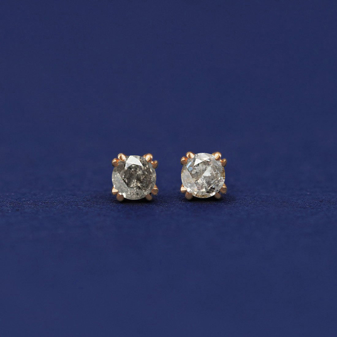 A pair of 14 karat gold stud earrings with 3 millimeter round Salt and Pepper diamonds on a dark blue background