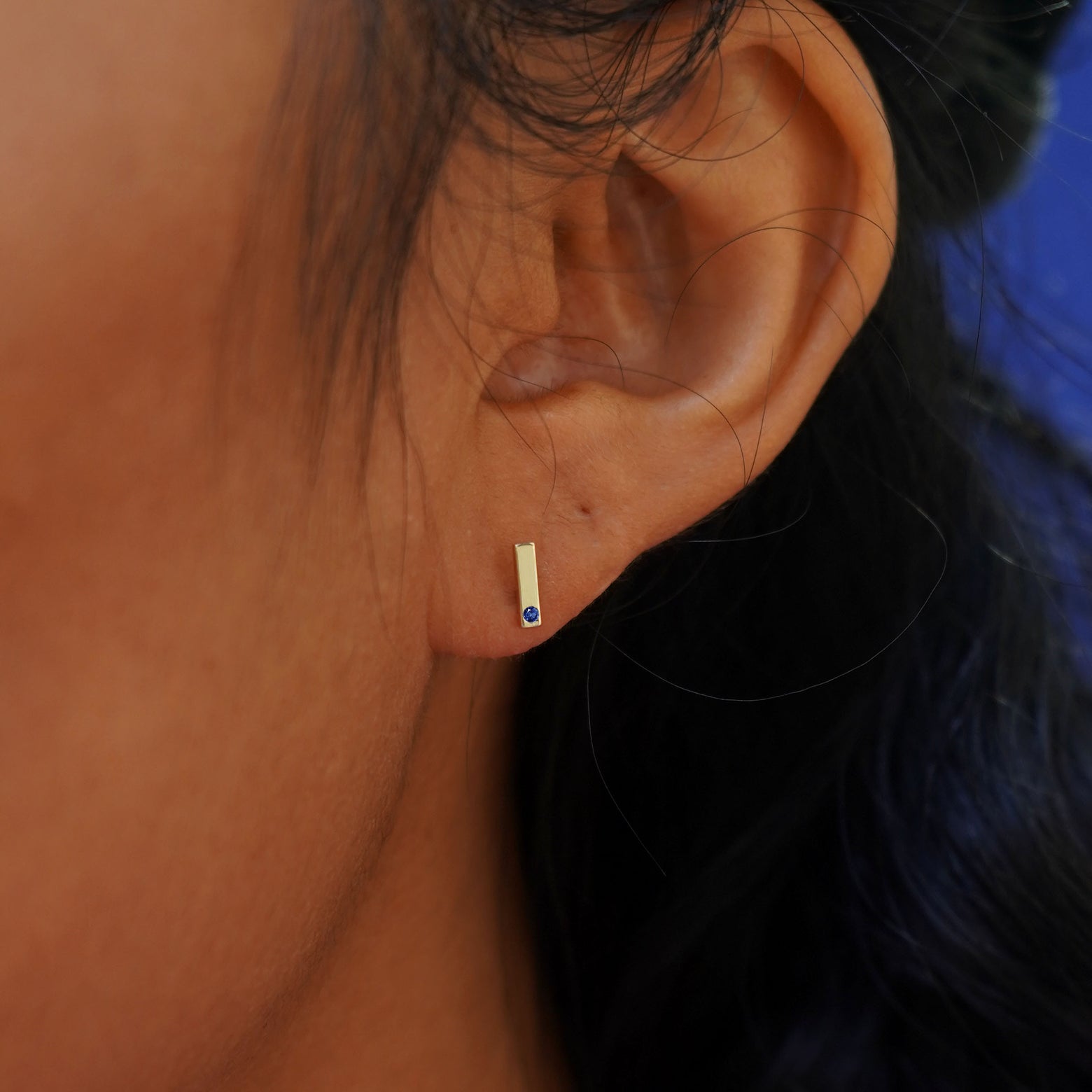 A model's ear wearing a 14k yellow gold blue sapphire Gemstone Bar Earring