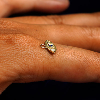 A 14k yellow gold blue sapphire Evil Eye Charm for earring balancing on the back of a model's finger