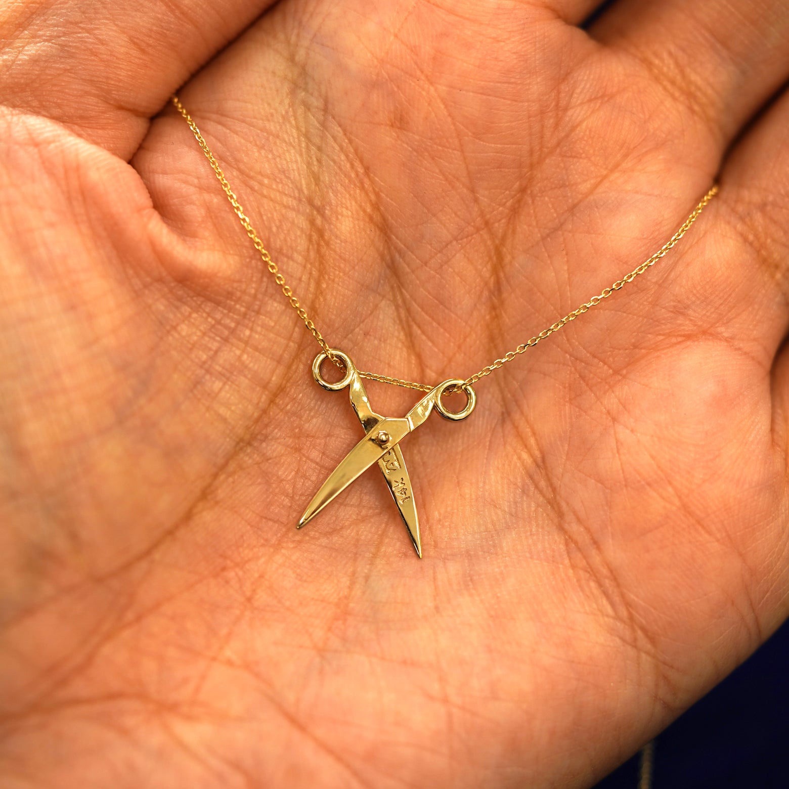 A yellow gold Scissors Necklace resting on a model's fingers with the scissors shown open to show the 14k and AU stamps