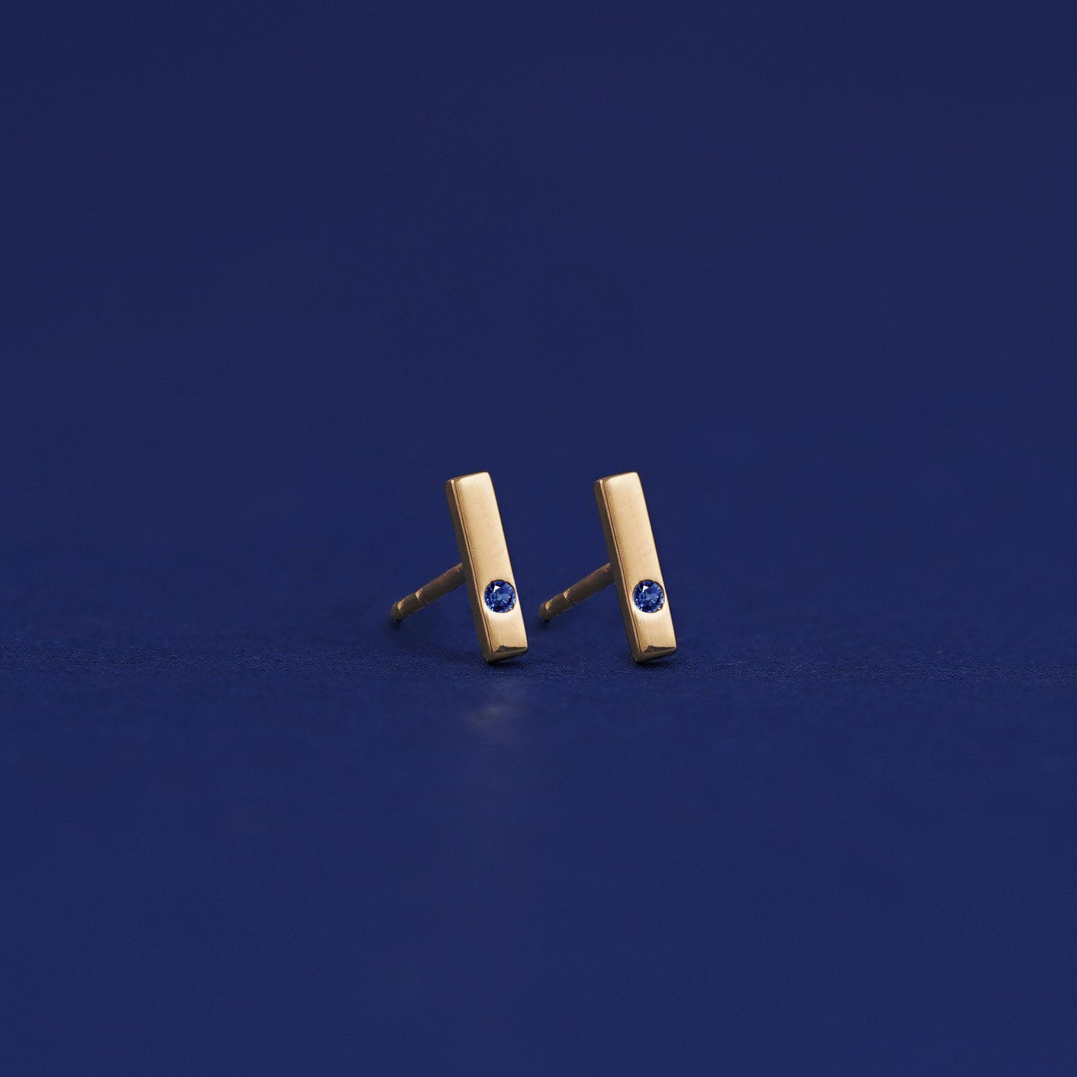 A pair of solid 14 karat yellow gold sapphire Gemstone Bar Earrings with no backings on a dark blue background