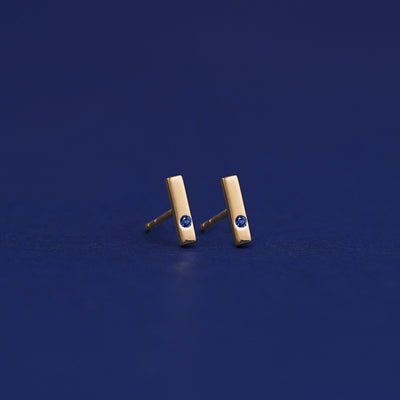 A pair of solid 14 karat yellow gold sapphire Gemstone Bar Earrings with no backings on a dark blue background