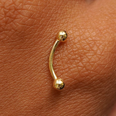 Close up view of a 14 karat gold Small Curved Barbell Piercing resting on the back of a model's hand