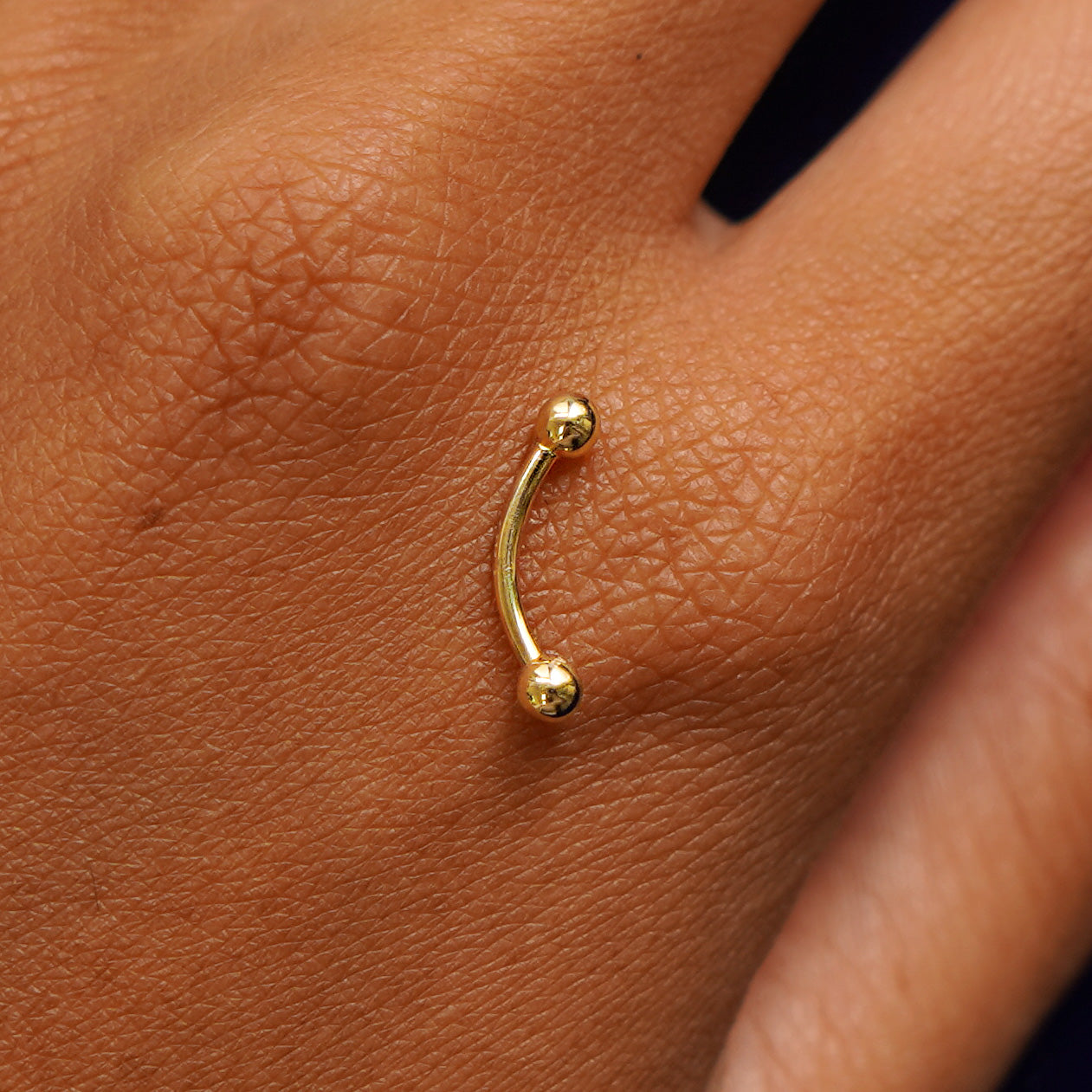 A yellow gold Small Curved Barbell Piercing resting on the back of a model's hand