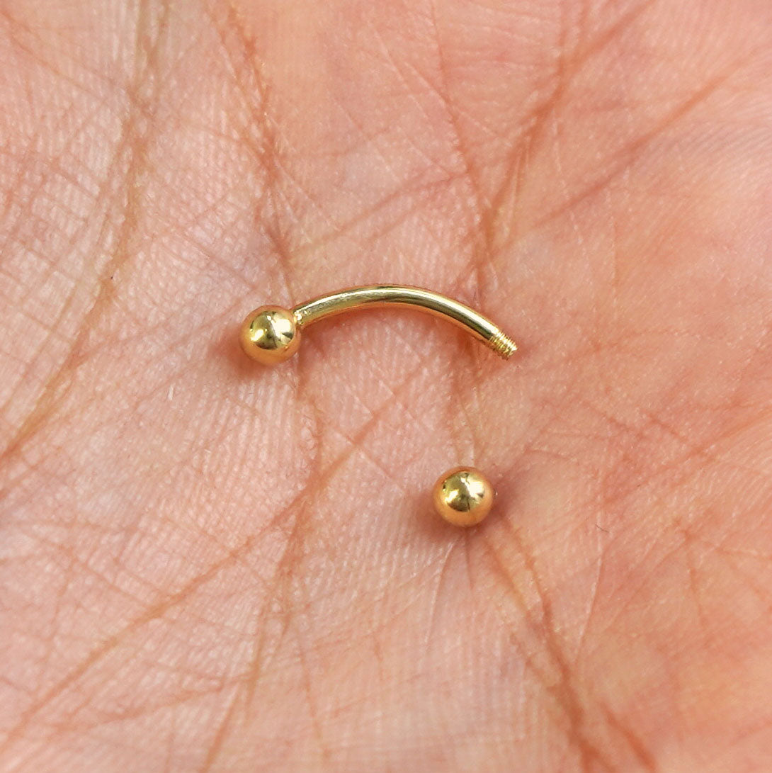 A yellow gold Small Curved Barbell Piercing with the externally threaded ball closure unscrewed in the palm of a model's hand