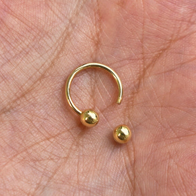 A solid yellow gold Small Horseshoe Piercing with the externally threaded ball closure unscrewed sitting in a model's palm
