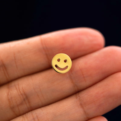 A solid 14k yellow gold Smiley Face Earring in between a model's fingers