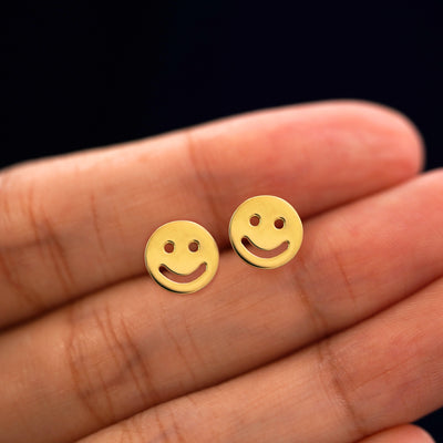 A model's hand holding a pair of recycled 14k gold Smiley Face Earrings