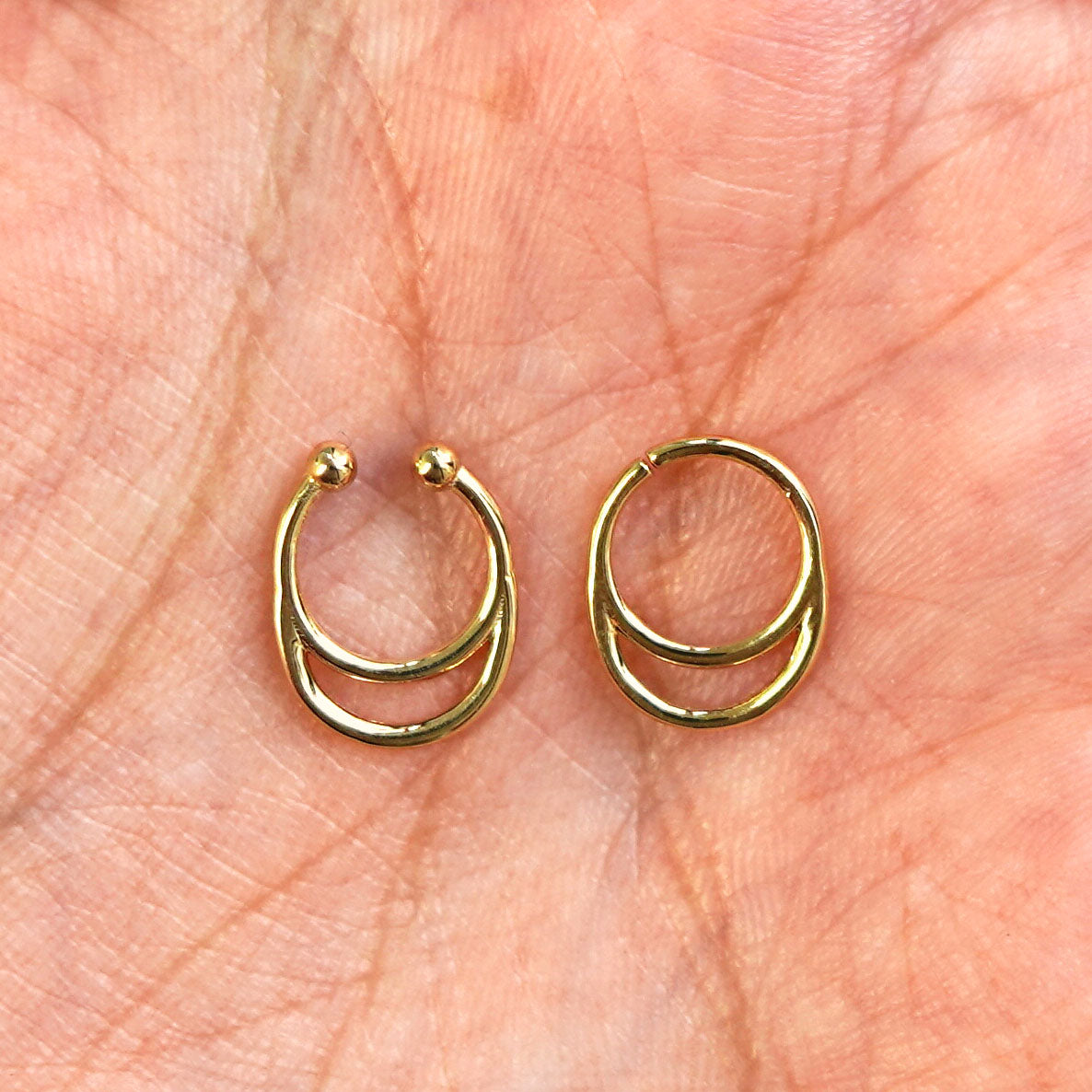 Two 14k gold Double Circle Septum rings show in options of Non-Pierced and Pierced resting in the palm of a model's hand