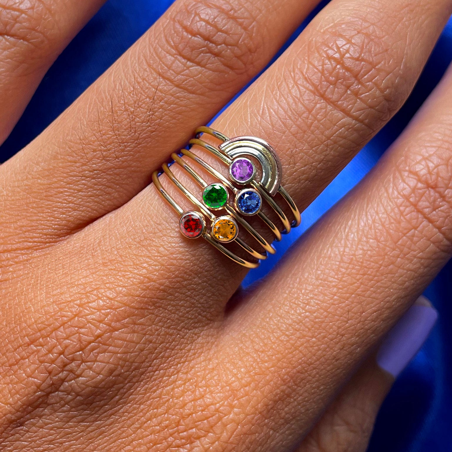 Close up view of a model's hand wearing a Rainbow Ring stacked above five gemstone rings of different colors