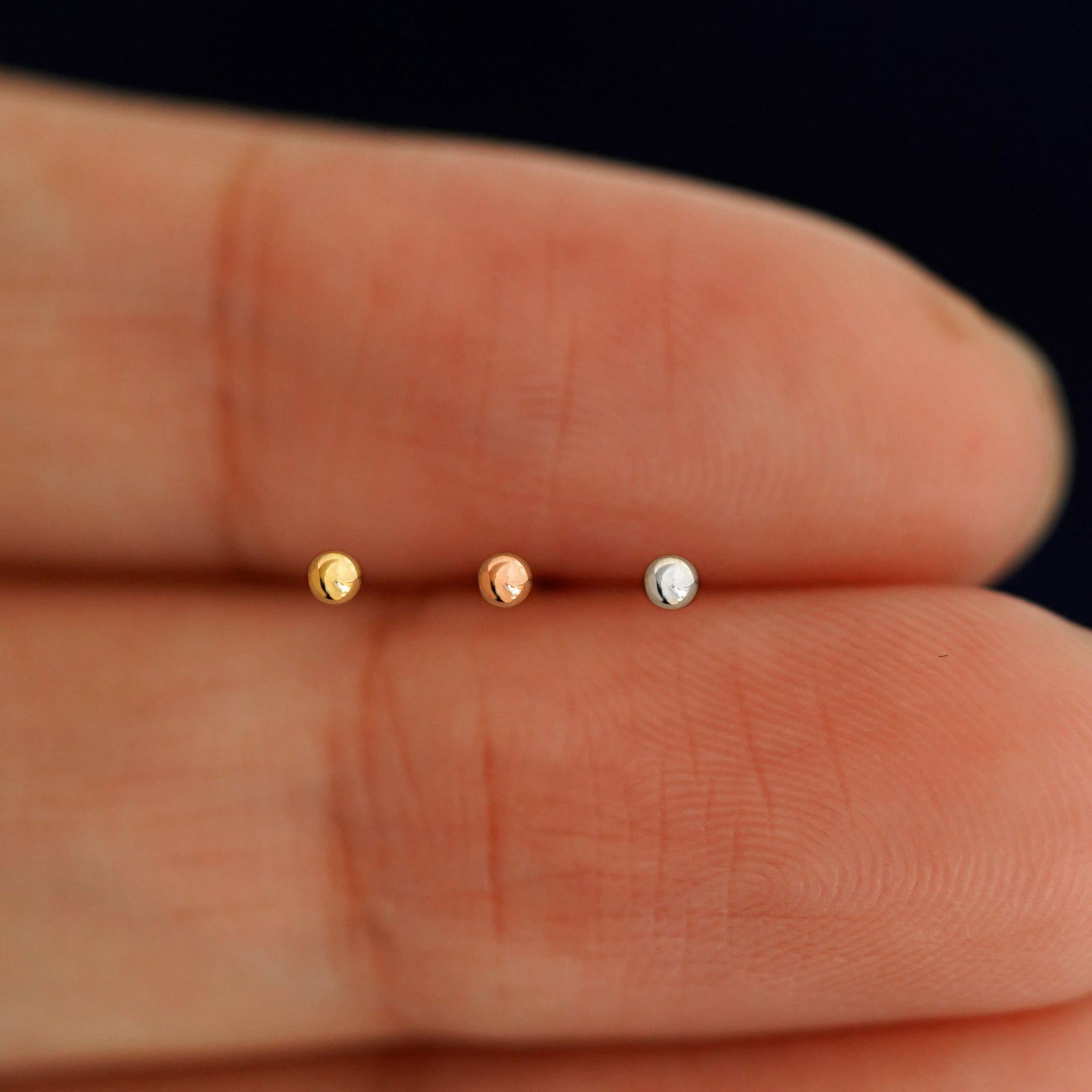 Three versions of the Mini Ball Earring showing options of yellow, rose, and white gold in between a model's fingers