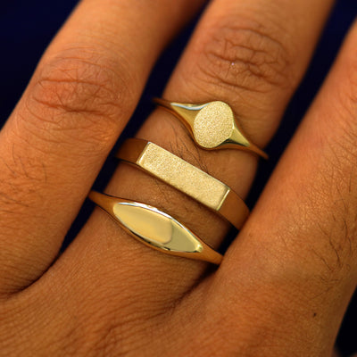 A model's fingers wearing a 14k yellow gold Rectangle, Oval, and Puffy Elongated Signet Rings stacked on their finger