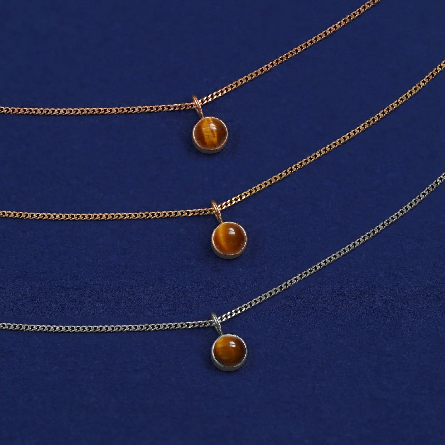 Three versions of the Tiger Eye Necklace in options of yellow, white, and rose gold on a dark blue background