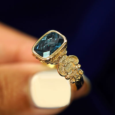 A model holding a solid yellow gold London blue topaz Royalty Ring tilted to show the details of scrollwork on the band