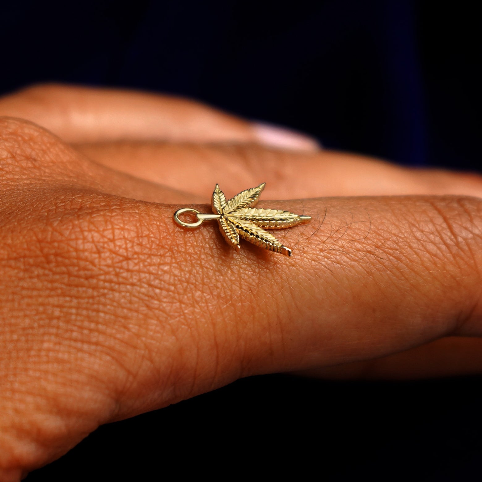 A 14k yellow gold Cannabis Charm for earring balancing on the back of a model's finger