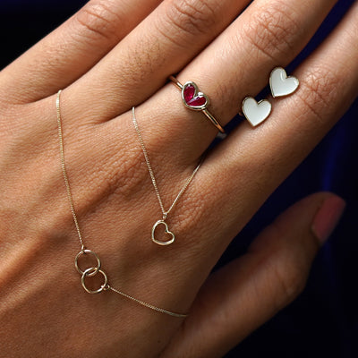 A hand holding a bound together necklace, a heart charm, and a pair of enamel heart earrings while wearing a ruby heart ring