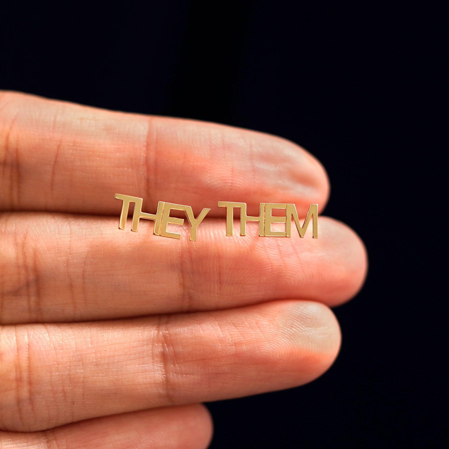 A pair 14k yellow gold They / Them Earring in between a model's fingers