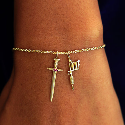 A model's wrist wearing a yellow gold Thick Cable Chain with a Sword Charm, and a Tattoo Machine Charm
