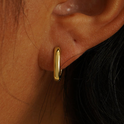 Close up view of a model's ear wearing a solid yellow gold Thick Oval Huggie Hoop