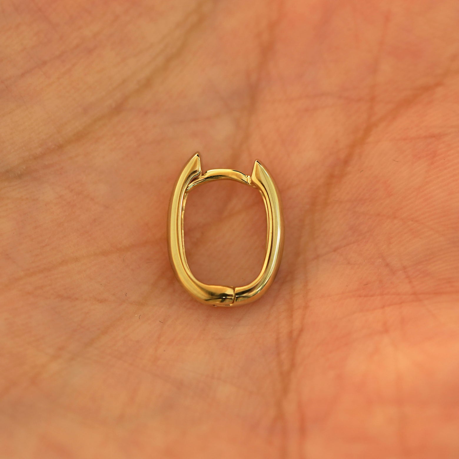 A single closed 14k yellow gold Thick Oval Huggie Hoop resting in a model's palm
