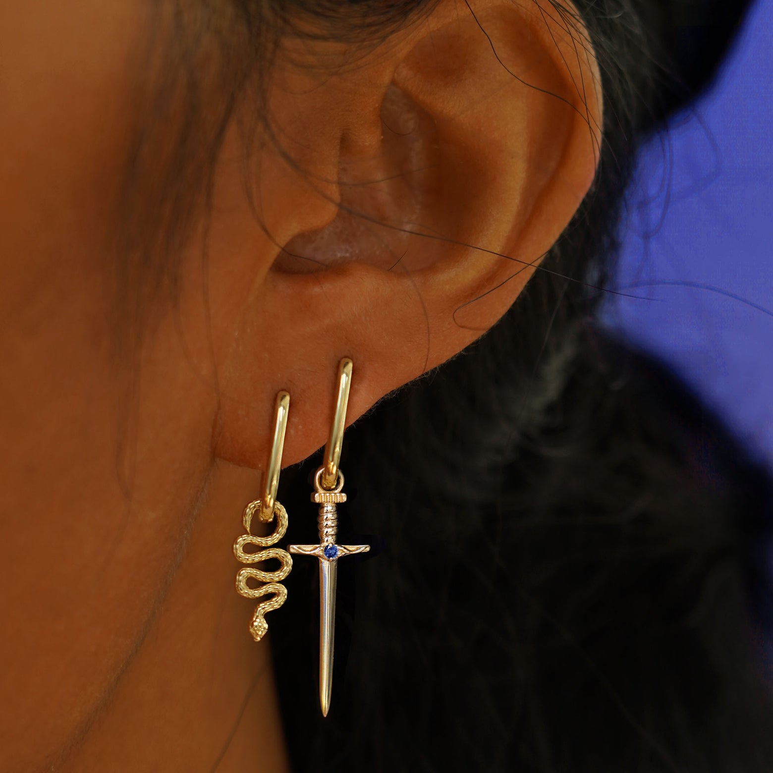 A model's ear wearing two Thin Oval Huggies one with a snake charm and one with a sword charm