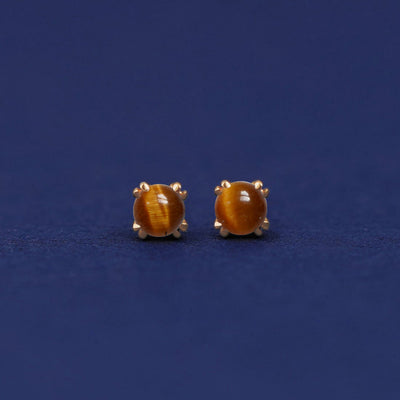 A pair of 14 karat gold stud earrings with 3 millimeter round Tiger Eyes on a dark blue background