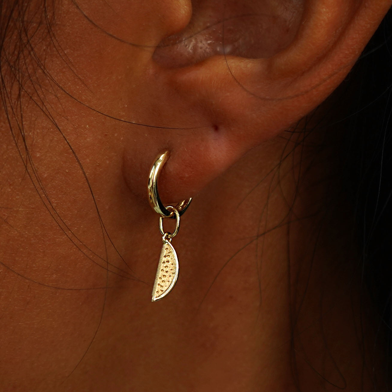 Close up view of a model's ear wearing a yellow gold Watermelon Charm on a Curvy Huggie Hoop