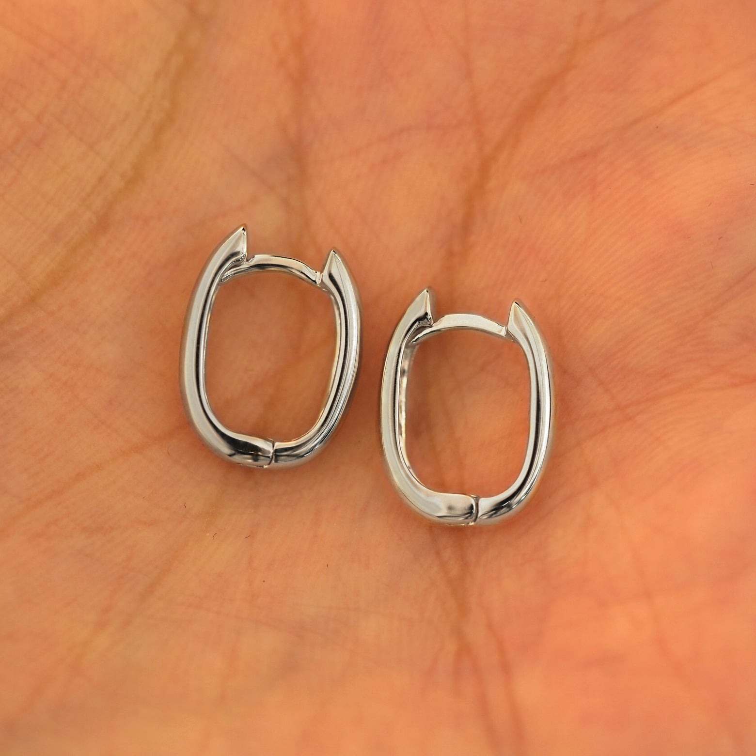 A pair of 14k white gold Thick Oval Huggie Hoops resting in a model's palm