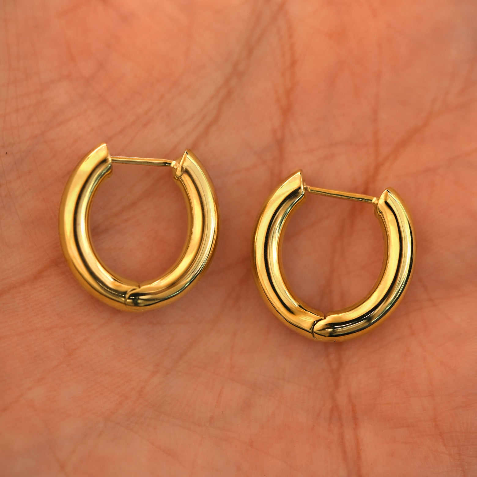 A pair of 14k yellow gold Chunky Oval Huggie Hoops resting in a model's palm