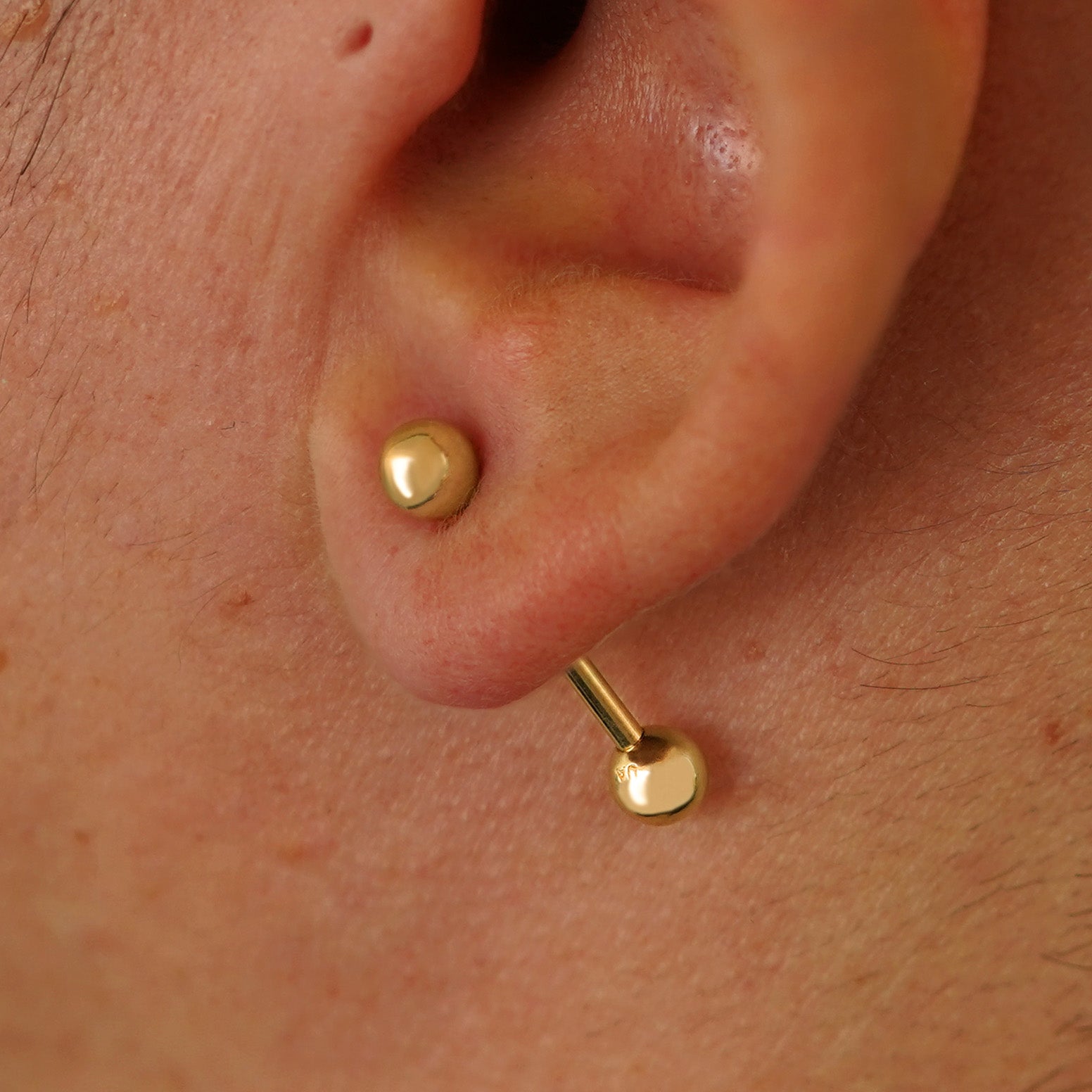 Close up view of a model's ear wearing a 14k yellow gold Medium Straight Barbell through a lobe piercing