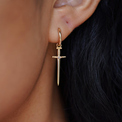 Close up view of a model's ear wearing a yellow gold Sword Charm on a Curvy Huggie Hoop