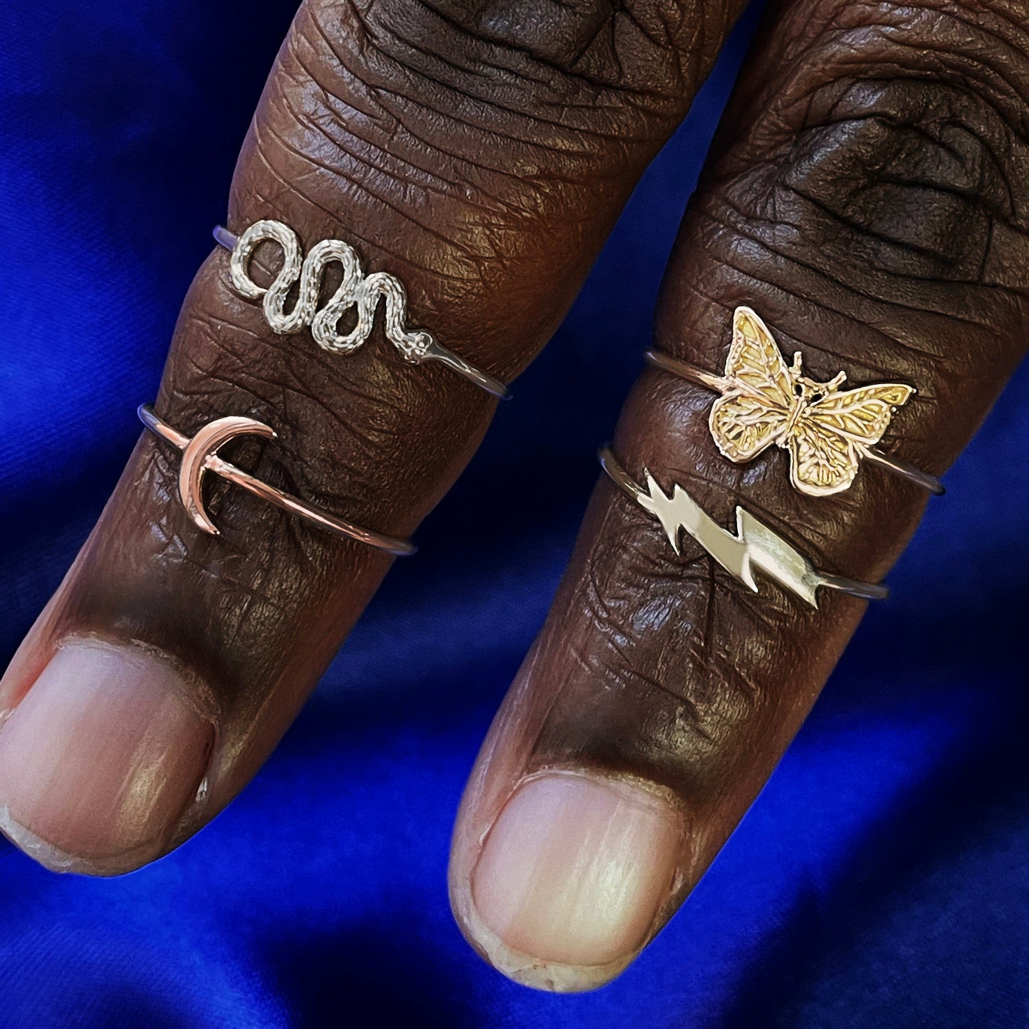 Two fingers wearings a white gold Snake Ring, rose gold Moon Ring, and yellow gold Lightning Bolt and Butterfly Rings