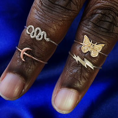 Two fingers wearings a white gold Snake Ring, rose gold Moon Ring, and yellow gold Lightning Bolt and Butterfly Rings