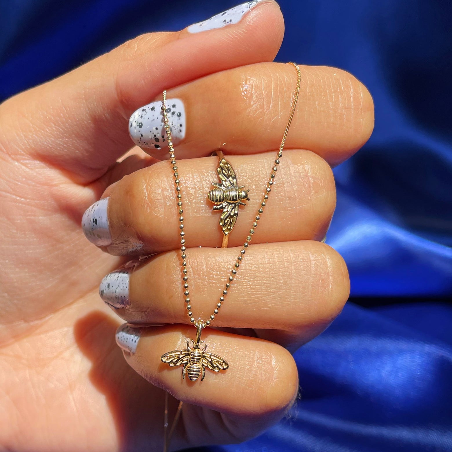 A model's hand wearing a 14k yellow gold bee ring while holding a Bead Chain necklace with a Bee Charm on it