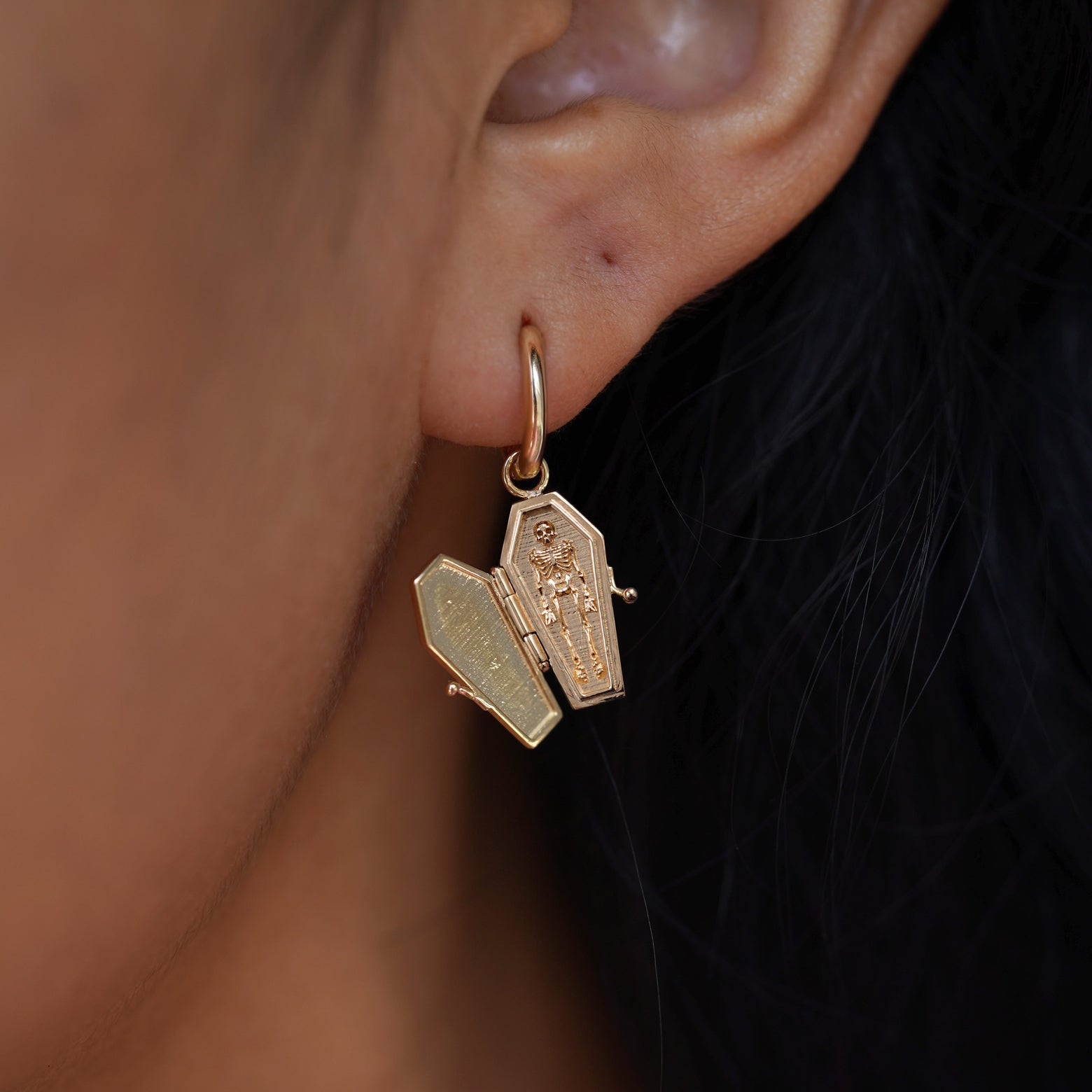 Close up view of a model's ear wearing a yellow gold Coffin Charm on a Curvy Huggie Hoop with the Coffin shown open