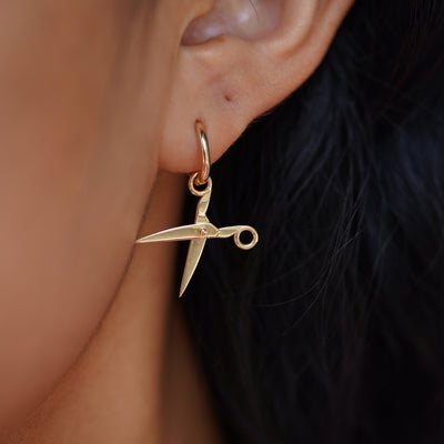 Close up view of a model's ear wearing a yellow gold Scissors Charm on a Curvy Huggie Hoop showing the scissors open