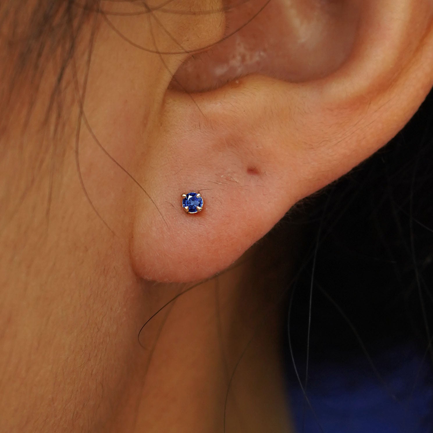 Close up view of a model's ear wearing a yellow gold 2mm Gemstone Flat Back Piercing in the option of sapphire