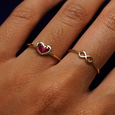 Close up view of a model's hand wearing an Infinity Ring on their middle finger and a Ruby Heart Ring on their ring finger