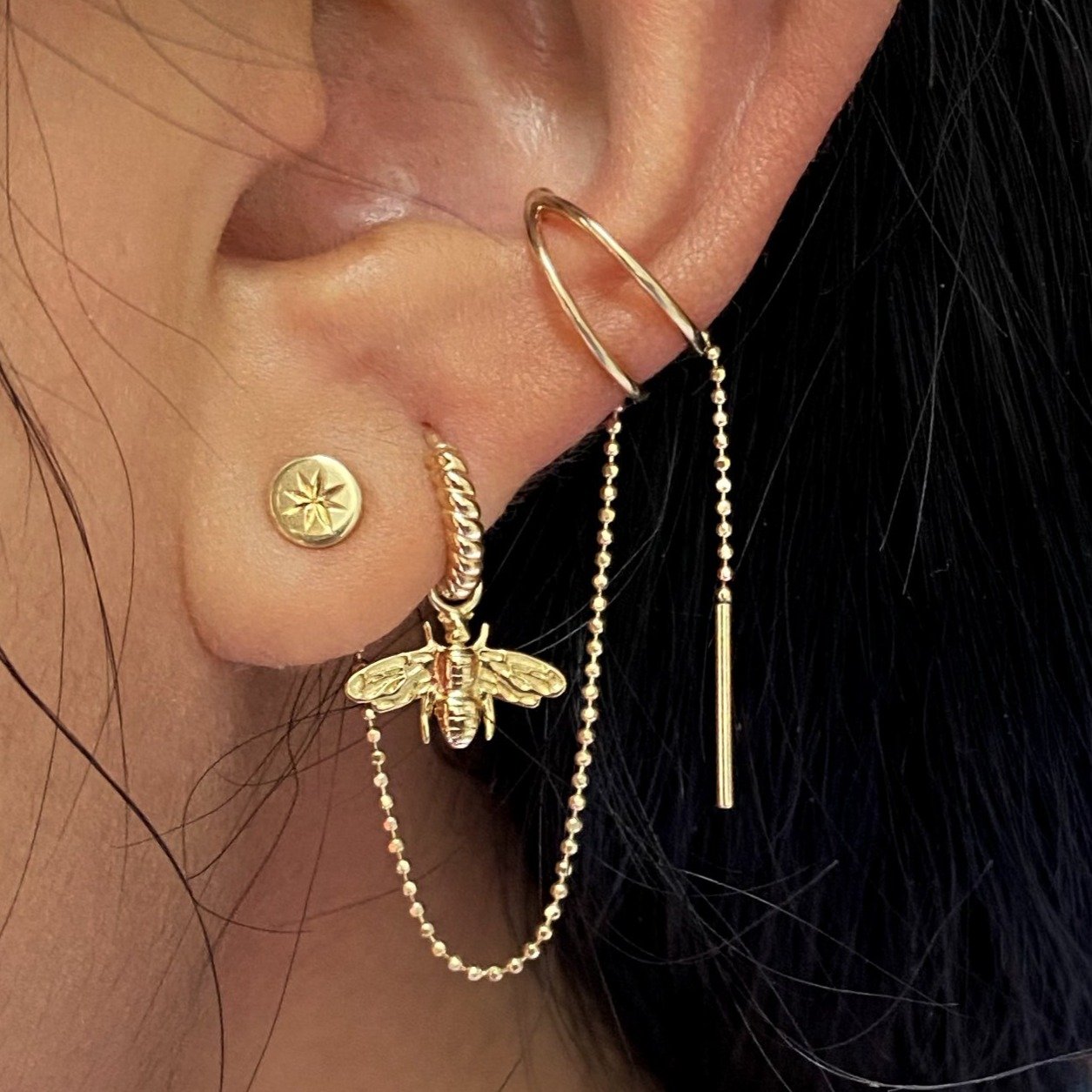 Close up view of a model's ear wearing, a yellow gold Bee Charm on a Rope Huggie Hoop and various Automic Gold earrings