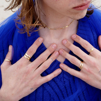 A model sitting on the beach with their hands touching their collarbone wearing various Automic Gold rings and necklaces