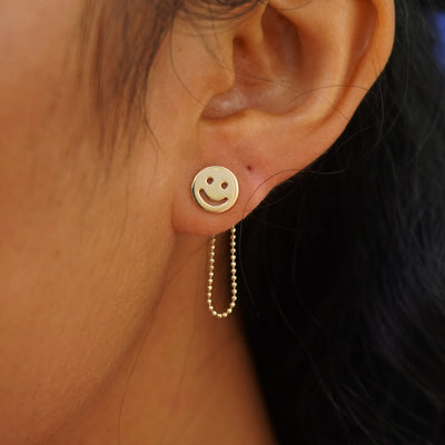 A model's ear wearing a yellow gold Connector with both attached behind a Smiley Face Earrings