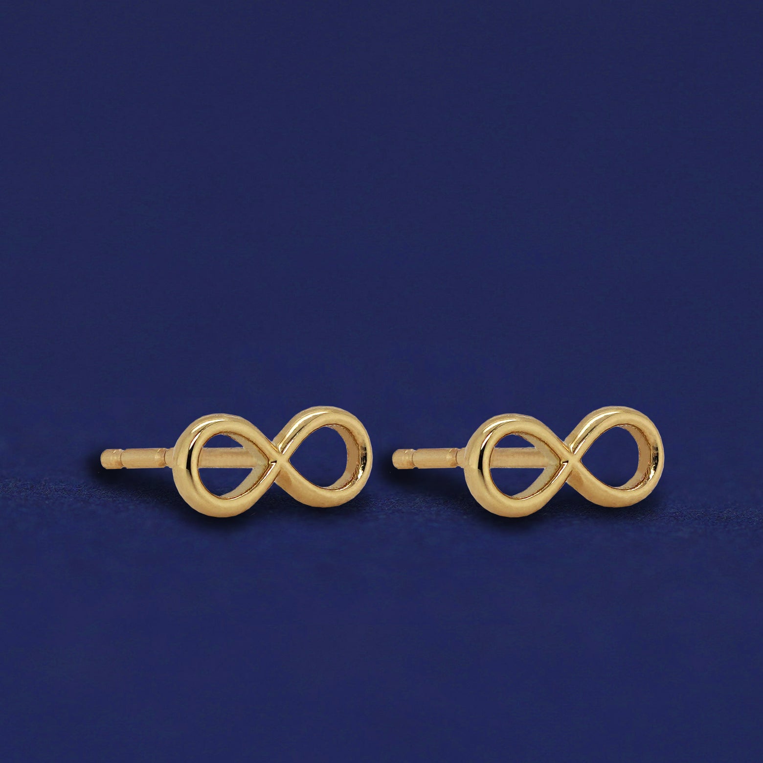 A pair of solid 14k gold Infinity Earrings on a dark blue background