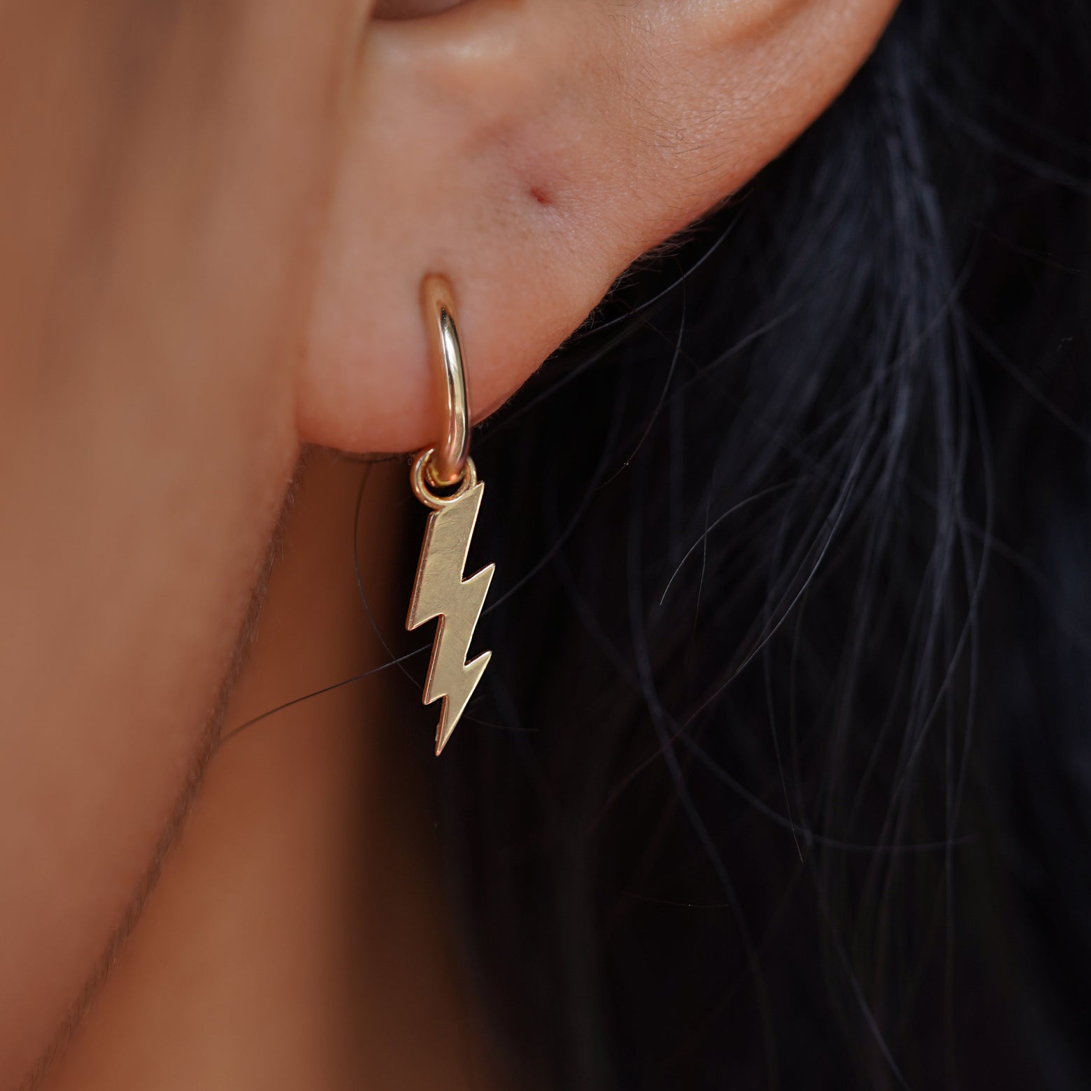 Close up view of a model's ear wearing a yellow gold Lightning Bolt Charm on a Curvy Huggie Hoop