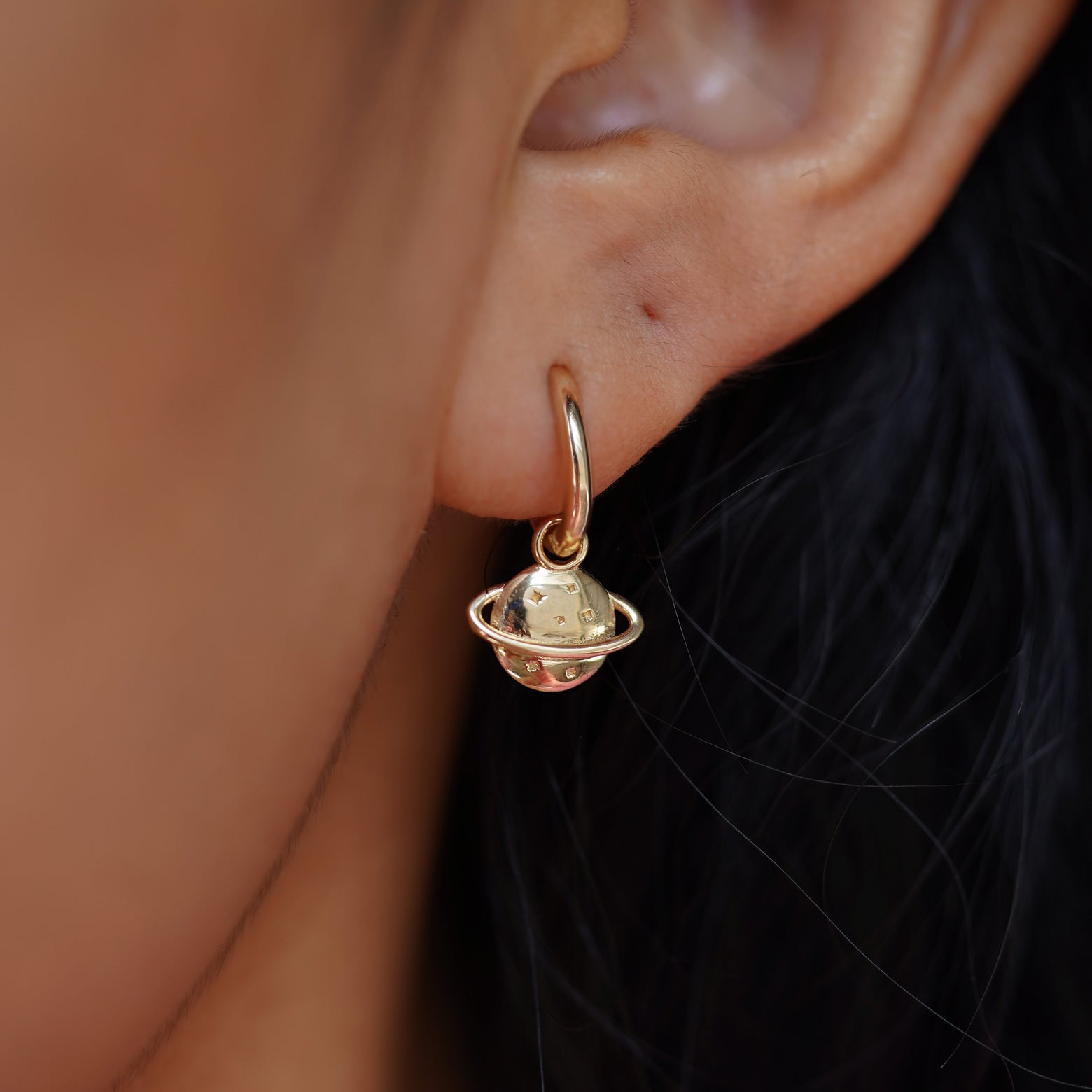 Close up view of a model's ear wearing a yellow gold Saturn Charm on a Curvy Huggie Hoop