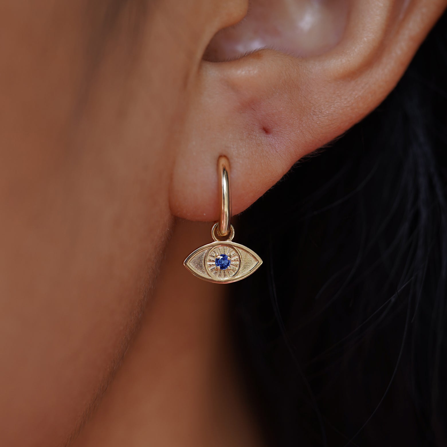 Close up view of a model's ear wearing a yellow gold sapphire Evil Eye Charm on a Curvy Huggie Hoop