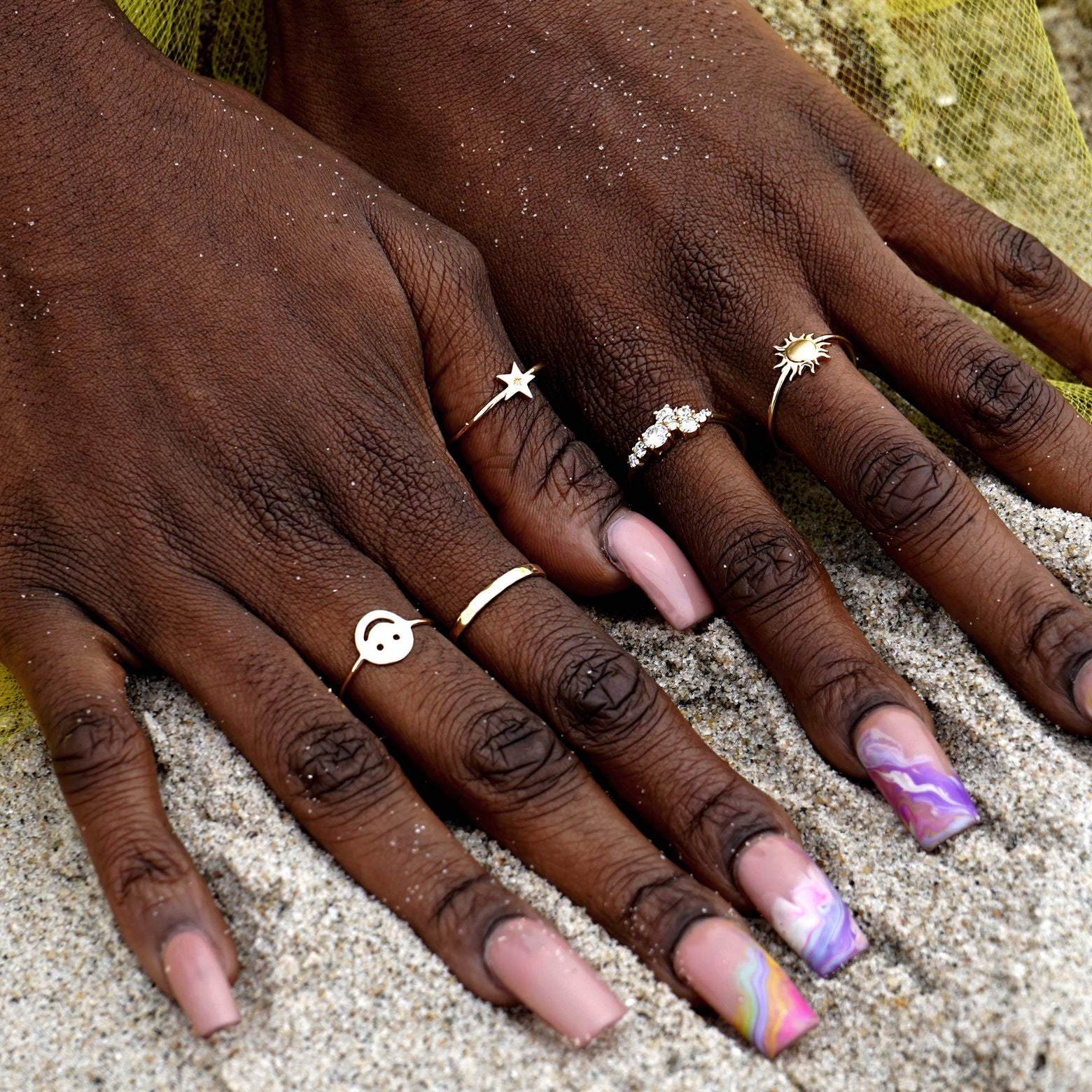 A model with long colorful nails digging their hands into the sand at the beach while wearing various Automic Gold Rings