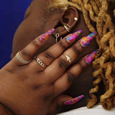 A model with long colorful nails touching the back of their head wearing various Automic Gold rings and earrings