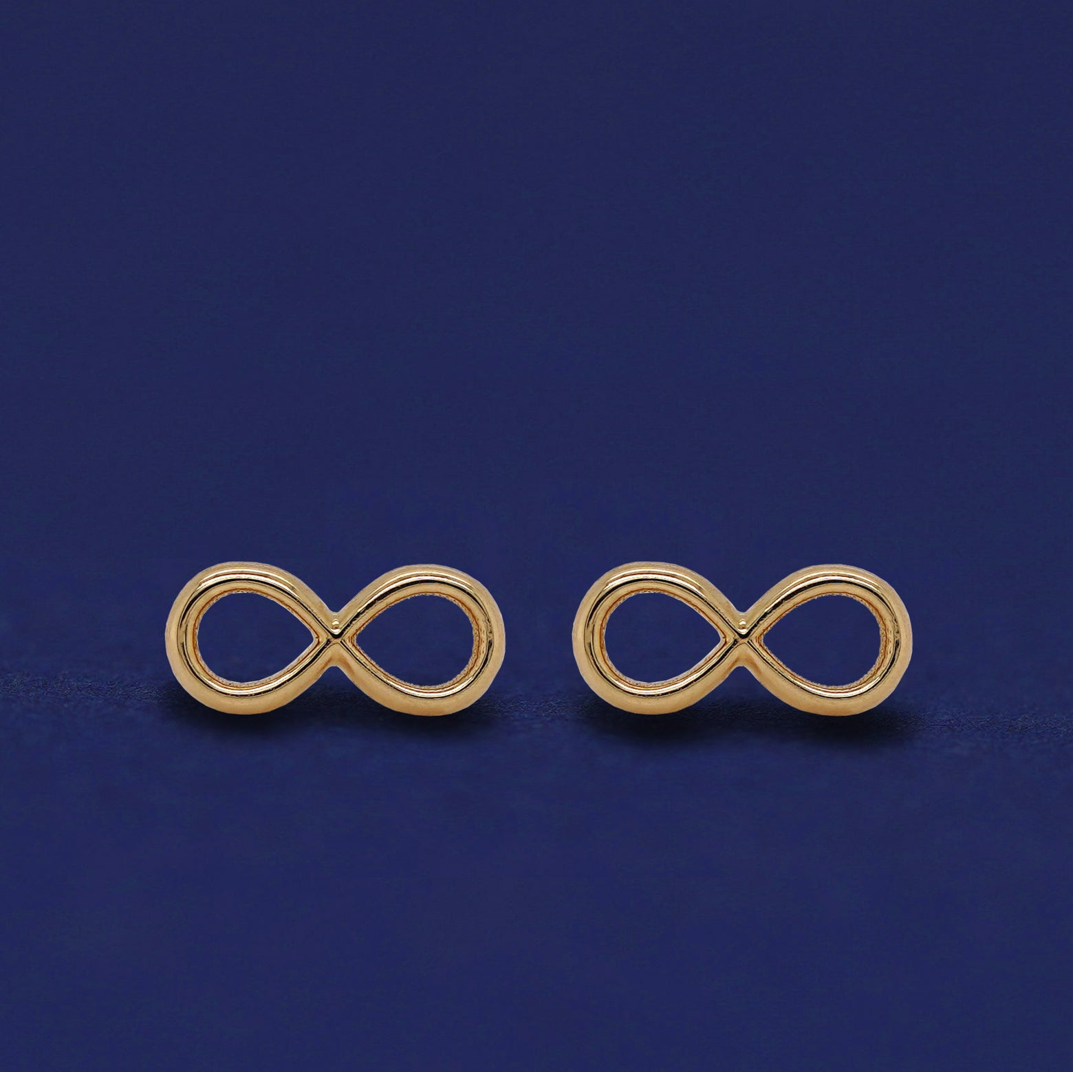 Front facing view of a pair of 14k yellow gold Infinity Earrings on a dark blue background