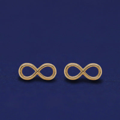 Front facing view of a pair of 14k yellow gold Infinity Earrings on a dark blue background